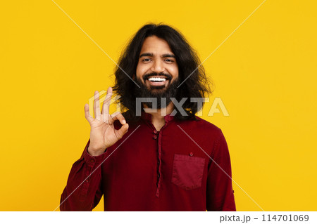 Indian man with a full beard and long hair smiles broadly, standing against a vivid yellow background, showing OK gesture with his right hand, expressing positivity and approval. Indian man with a full beard and long hair smiles broadly, standing against a vivid yellow background, showing OK gesture with his right hand, expressing positivity and approval. 114701069