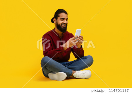 Indian man is seated on the floor, engrossed in his cell phone. He appears focused as he scrolls through the screen, possibly engaging in messaging or browsing Indian man is seated on the floor, engrossed in his cell phone. He appears focused as he scrolls through the screen, possibly engaging in messaging or browsing 114701157