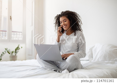 Hispanic woman is sitting on a bed, multitasking by talking on her cell phone while using a laptop. She appears focused and engaged in her activities, balancing both devices comfortably. Hispanic woman is sitting on a bed, multitasking by talking on her cell phone while using a laptop. She appears focused and engaged in her activities, balancing both devices comfortably. 114701161