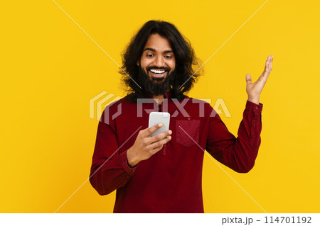 Indian man with long hair and a beard is seen holding a cell phone in his hand. The man appears focused on the screen, possibly texting or browsing, raising hand up 114701192