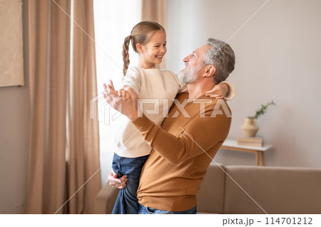 Senior man is standing, holding a smiling little girl in his arms. They both appear happy and connected, showcasing a heartwarming moment of familial love. 114701212