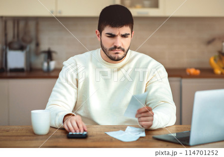 A man is depicted sitting at a table with a calculator in front of him, appearing to be engaged in calculations or financial tasks. He is focused on the calculator screen A man is depicted sitting at a table with a calculator in front of him, appearing to be engaged in calculations or financial tasks. He is focused on the calculator screen 114701252