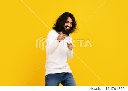 Indian man with long hair and a beard is standing in front of a bright yellow background, pointing at camera and smiling 114701253