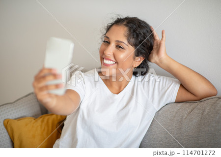 A woman is seated on a couch, holding a cell phone in her hands. She appears focused on the screen, engrossed in whatever she is viewing or doing on the device, taking selfie 114701272