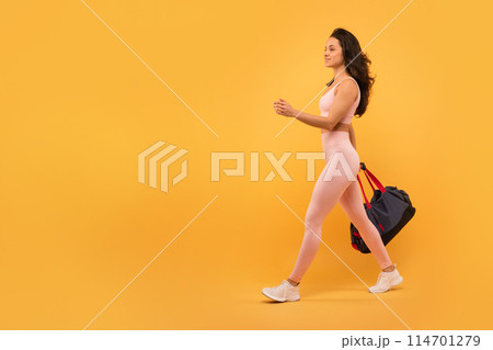 A young woman with long hair, dressed in a form-fitting activewear outfit, walks with purpose while carrying a gym bag, isolated on yellow background 114701279