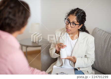 A woman in a white coat is seated across from a patient, engaging in a detailed explanation about a medication. She holds a pill bottle in her hand and gestures to signify the directions for use A woman in a white coat is seated across from a patient, engaging in a detailed explanation about a medication. She holds a pill bottle in her hand and gestures to signify the directions for use 114701299