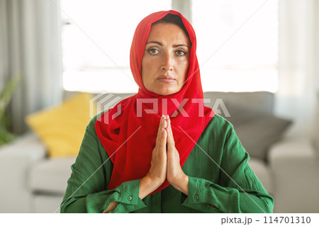 A woman is depicted wearing a red and green hijab. The hijab covers her head and neck, with the colors contrasting against each other, praying at home 114701310
