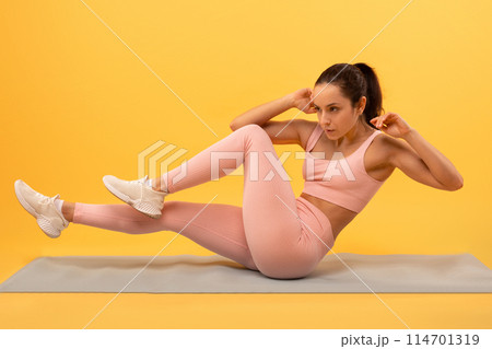 A focused young woman in a pink athletic outfit is engaged in a fitness routine, executing bicycle crunches on a blue yoga mat, vivid yellow backdrop that highlights her exercise activity. 114701319