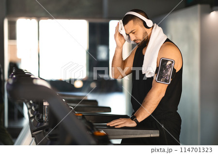 A focused young man with a towel around his neck is taking a break after running on a treadmill. He is wearing a black sleeveless shirt and has a smartphone strapped to his arm 114701323