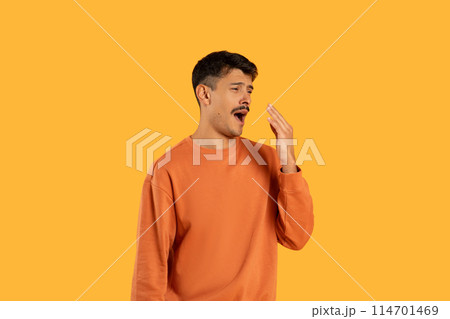 A young man with dark hair stands against a vivid yellow backdrop, covering his mouth with his hand as he yawns widely, displaying signs of fatigue or boredom wearing a casual orange sweatshirt. 114701469