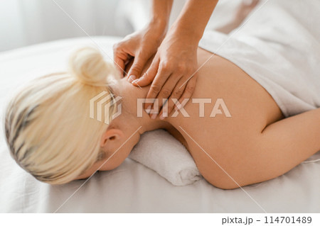 Client woman with blonde hair receives a deep tissue massage from a skilled therapist. She is lying face down on a massage table, her head resting comfortably on a face cradle. 114701489