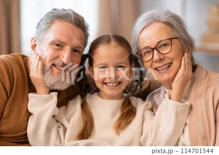 A warm and affectionate moment as a grey-haired grandfather and grandmother sit closely with their beaming young granddaughter, looking directly at the camera A warm and affectionate moment as a grey-haired grandfather and grandmother sit closely with their beaming young granddaughter, looking directly at the camera 114701544