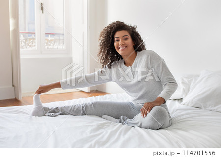 A cheerful Hispanic woman with curly hair is sitting on a neatly made bed in a bright bedroom. She is wearing a cozy white sweater and gray pants A cheerful Hispanic woman with curly hair is sitting on a neatly made bed in a bright bedroom. She is wearing a cozy white sweater and gray pants 114701556