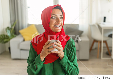A cheerful woman wearing a red hijab and green blouse stands in a sunlit living room, holding a gray mug with both hands. She gazes out of the frame with a joyful expression A cheerful woman wearing a red hijab and green blouse stands in a sunlit living room, holding a gray mug with both hands. She gazes out of the frame with a joyful expression 114701563