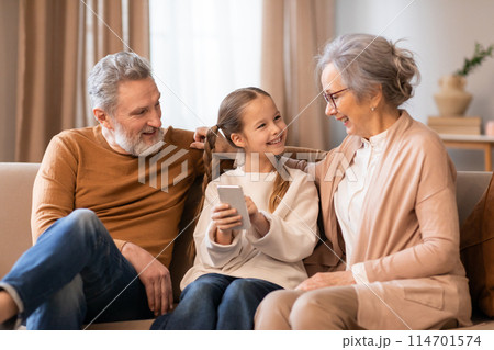 A family of three individuals, consisting of grandparents and little girl, sitting together on a couch in a living room. They are focused on a cell phone screen A family of three individuals, consisting of grandparents and little girl, sitting together on a couch in a living room. They are focused on a cell phone screen 114701574