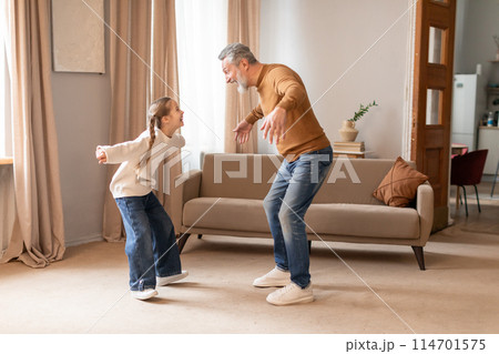 A heartwarming scene unfolds as a grandfather and his young granddaughter engage in a playful dance, with smiles on their faces, in the comfort of a warm, sunlit living room. A heartwarming scene unfolds as a grandfather and his young granddaughter engage in a playful dance, with smiles on their faces, in the comfort of a warm, sunlit living room. 114701575