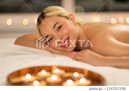 Smiling stunning woman is lying gracefully on top of a bed, placed next to flickering candles. The soft glow of the candles illuminates her features as she relaxes in this cozy setting. Smiling stunning woman is lying gracefully on top of a bed, placed next to flickering candles. The soft glow of the candles illuminates her features as she relaxes in this cozy setting. 114701586