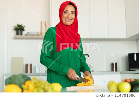 A woman wearing a red and green hijab is skillfully cutting a bell-pepper on a wooden cutting board. She is focused on her task, delicately slicing through the cheese with precision. 114701600