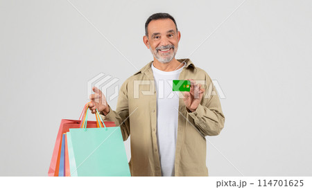 A senior man standing holding shopping bags in one hand and a credit card in the other. He appears to be in a shopping area, possibly a mall, ready to make purchases. A senior man standing holding shopping bags in one hand and a credit card in the other. He appears to be in a shopping area, possibly a mall, ready to make purchases. 114701625
