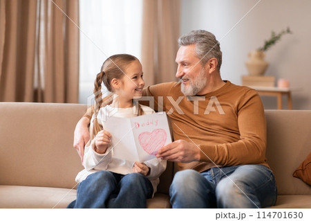 Senior man and a little girl are seated on a couch in a living room. Grandfather appears to be talking to the girl, who is looking attentively at him, engaged in conversation or bonding time. 114701630