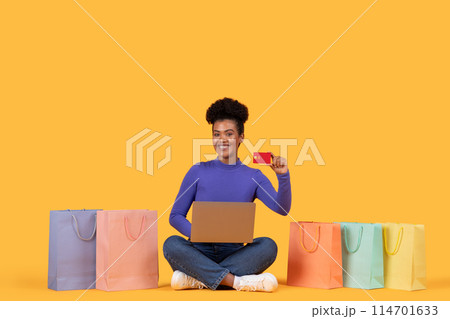 A cheerful young Hispanic woman sits cross-legged between colorful shopping bags, proudly displaying a credit card while looking at an open laptop, suggesting a successful online shopping session A cheerful young Hispanic woman sits cross-legged between colorful shopping bags, proudly displaying a credit card while looking at an open laptop, suggesting a successful online shopping session 114701633