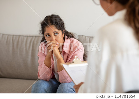 A young woman sits on a couch, her hands clasped together in a gesture of concern, as she engages in a therapy session. A therapist, partially visible, sits across from her with a clipboard 114701654