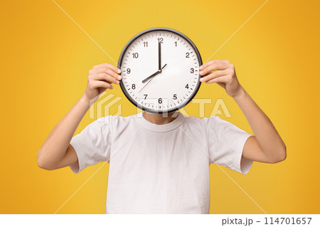 Time management concept. Teen boy covering his face with big clock, orange panorama background 114701657