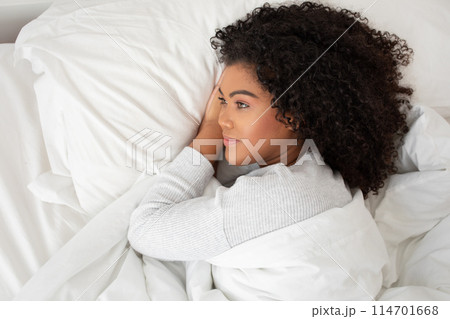 Hispanic woman is lying down in bed, covered by a white blanket. She appears relaxed and comfortable, with her eyes closed as if resting or sleeping peacefully. 114701668