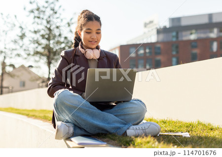 A girl is sitting on a bench, engrossed in her laptop computer. She appears focused and concentrated as she types on the keyboard. 114701676