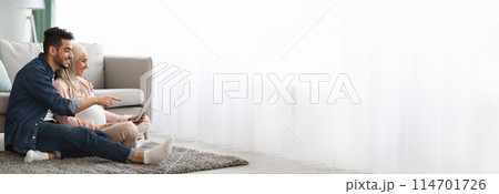 Middle eastern pregnant couple lounges comfortably while using a tablet together, with plenty of copy space for web-banner design on the side 114701726
