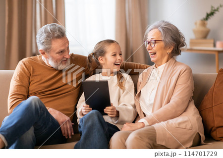 A young girl, seated between her grandparents, shares a joyful moment with them on a comfortable couch, using digital tablet together at home A young girl, seated between her grandparents, shares a joyful moment with them on a comfortable couch, using digital tablet together at home 114701729