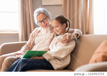 An older woman and a young girl are seated on a couch at home, engaging in conversation while reading book together An older woman and a young girl are seated on a couch at home, engaging in conversation while reading book together 114701755