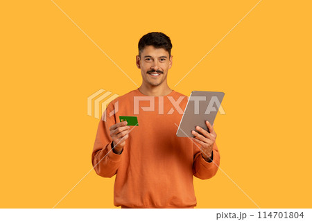 A man is shown in the image holding a tablet in one hand and a credit card in the other. He appears to be making a purchase or conducting online banking using the digital device and the card. A man is shown in the image holding a tablet in one hand and a credit card in the other. He appears to be making a purchase or conducting online banking using the digital device and the card. 114701804