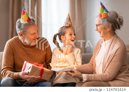 A young girl, wearing a festive party hat, gleefully receives a birthday cake from her grandmother, while an equally joyous grandfather looks on holding a gift 114701814
