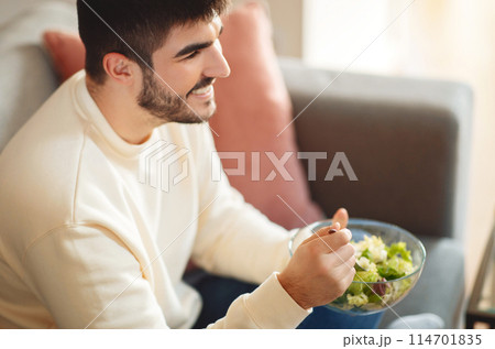 A man is seated on a couch, holding a bowl of food in his hands. He looks relaxed as he enjoys his meal in the comfort of his living room. 114701835