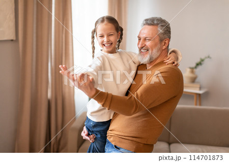 Senior man is standing, holding a little girl in his arms, grandfather dancing with his cute little granddaughter at home Senior man is standing, holding a little girl in his arms, grandfather dancing with his cute little granddaughter at home 114701873