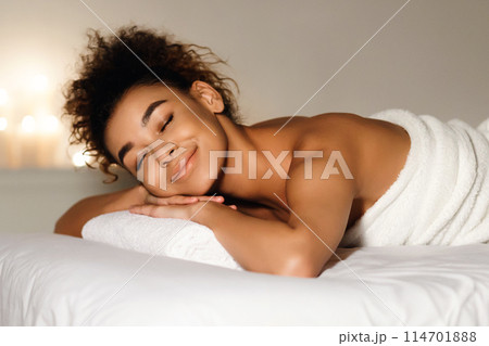 Close-up of a joyful african american woman with a radiant smile, wrapped in a towel in a spa environment 114701888