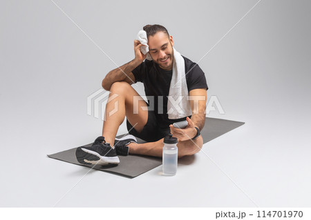 A man is seated on a floor mat with a towel draped around his neck. He appears relaxed and may have just finished exercising or working out, checking smartphone A man is seated on a floor mat with a towel draped around his neck. He appears relaxed and may have just finished exercising or working out, checking smartphone 114701970