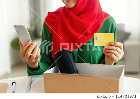 A woman in hijab is seated at a table, holding a smart phone in one hand and a credit card in the other. She appears to be engaged with the device, making a purchase or checking her finances, cropped 114701971