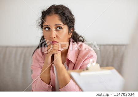 A young woman with curly hair is sitting at a table in a room with soft natural light filtering in, listens to her therapist during session at clinic 114702034