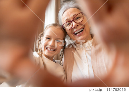 An older woman and a young girl are smiling and holding a phone together to take a selfie. They are standing close to each other and seem to be enjoying the moment. 114702056
