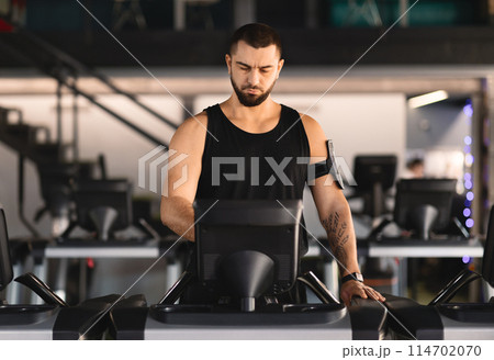 A man wearing athletic clothing is jogging on a treadmill machine inside a bright and modern gym. The equipment is surrounded by other exercise machines and gym-goers in the background. 114702070