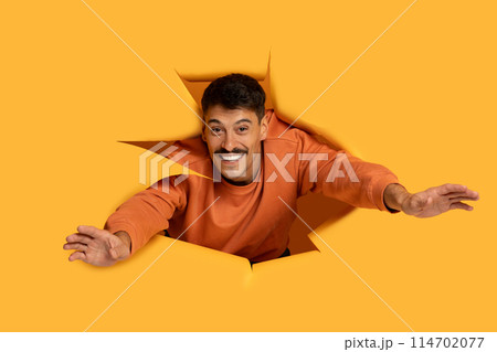 A man wearing an orange shirt is forcefully breaking through a bright yellow wall. His hands and arms are pushing against the wall as debris scatters around him. 114702077