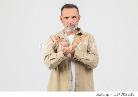 A senior man is shown standing upright with his hands folded in front of his chest in a confident and attentive posture, showing stop gesture on white 114702139
