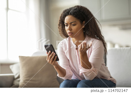 This image shows an African American woman on her couch, frowning at her phone, surrounded by a comfortable home environment, depicting a moment of stress This image shows an African American woman on her couch, frowning at her phone, surrounded by a comfortable home environment, depicting a moment of stress 114702147
