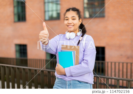 A young girl wearing headphones is seen holding a book and giving a thumbs up gesture. She appears engaged in reading while listening to music, showing approval with her hand signal. 114702156