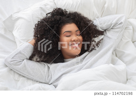 Hispanic woman is lying in bed with her head resting on her hands, looking contemplative and relaxed in the morning. The room is dimly lit, emphasizing her peaceful expression. 114702198
