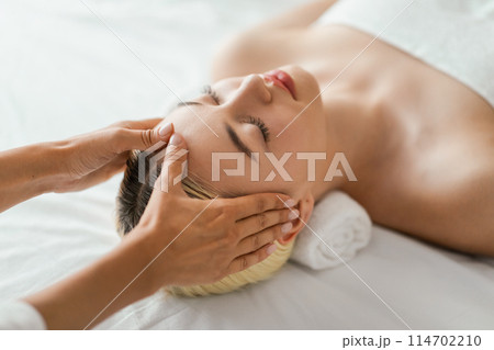A woman is lying down with her eyes closed, receiving a relaxing facial massage. The therapists hands are gently massaging her face, view above A woman is lying down with her eyes closed, receiving a relaxing facial massage. The therapists hands are gently massaging her face, view above 114702210