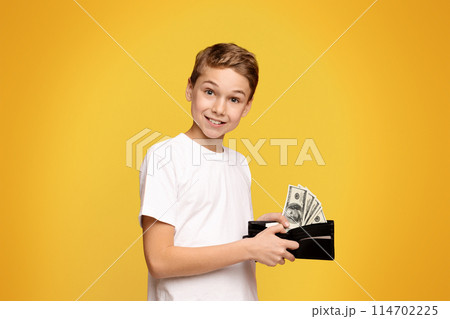 Rich teenager. Happy little boy holding wallet with lots of dollars, orange studio background Rich teenager. Happy little boy holding wallet with lots of dollars, orange studio background 114702225