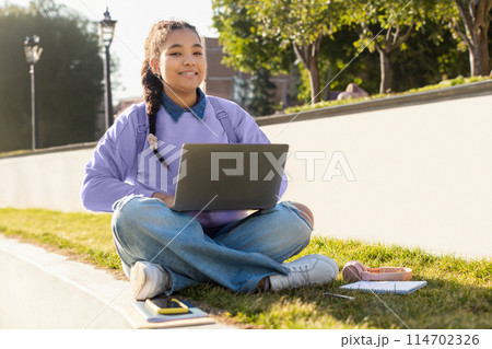 A girl student is sitting on the grass outdoors, working on her laptop. She appears focused and engaged in her work, surrounded by greenery, copy space 114702326